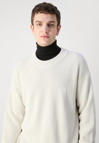 Maglione a coste color crema con scollatura rotonda, caratterizzato da una consistenza morbida e un piccolo patch con logo. Indossato sopra un dolcevita nero.