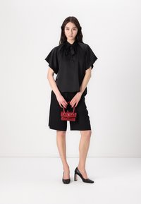 Top nero a maniche corte con collo a legaccio, abbinato a pantaloni shorts neri. Il modello tiene in mano una piccola borsa rossa con dettagli in argento e indossa tacchi neri.