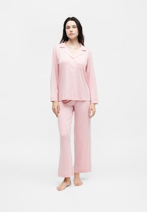 Γυναίκα στέκεται ξυπόλητη φορώντας ροζ ανοιχτό ροζ πυτζάμα με μακριά μανίκια και κουμπιά και αντίστοιχα φαρδιά πυτζάμα παντελόνια σε απλό λευκό φόντο.