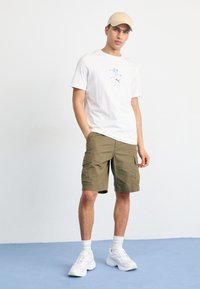 Camiseta de algodón blanca con gráfico azul, pantalones cortos cargo verde oliva, zapatillas deportivas blancas y una gorra beige. Conjunto simple y casual contra un fondo plano.