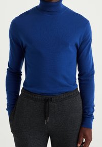 Blå polo-tröja i ett slätt stickat material med långa ärmar. Bärs med mörkgrå byxor med dragsko som har ett texturerat mönster.