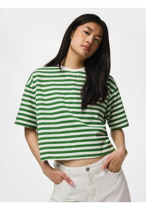 Jeune femme aux longs cheveux foncés portant un t-shirt à manches courtes rayé vert et blanc et un pantalon blanc, les mains dans les poches, expression neutre.