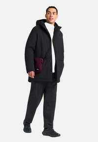 Veste imperméable noire avec capuche, associée à un pantalon assorti et des baskets noires. Un petit sac crossbody violet est porté en bandoulière.