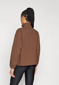 Καφέ φαρδιά ζακέτα με ψηλό γιακά, ελαστικά μανίκια και χαλαρή εφαρμογή. Λείο ύφασμα με ελάχιστες ραφές.