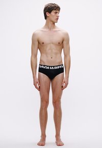 Μαύρο εσώρουχο τύπου σλιπ με φαρδιά λευκή ζώνη μέσης με λογότυπο. Το μοντέλο στέκεται όρθιο με ουδέτερη έκφραση μπροστά σε απλό φόντο.