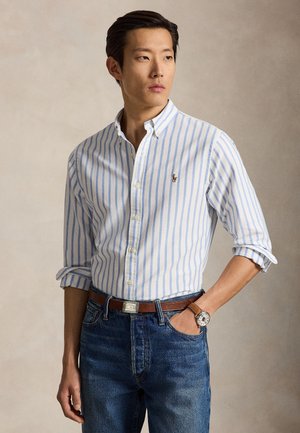 Homme portant une chemise rayée blanche et bleue avec des manches retroussées, un jean bleu, une ceinture marron et une montre-bracelet, debout avec une main dans la poche.