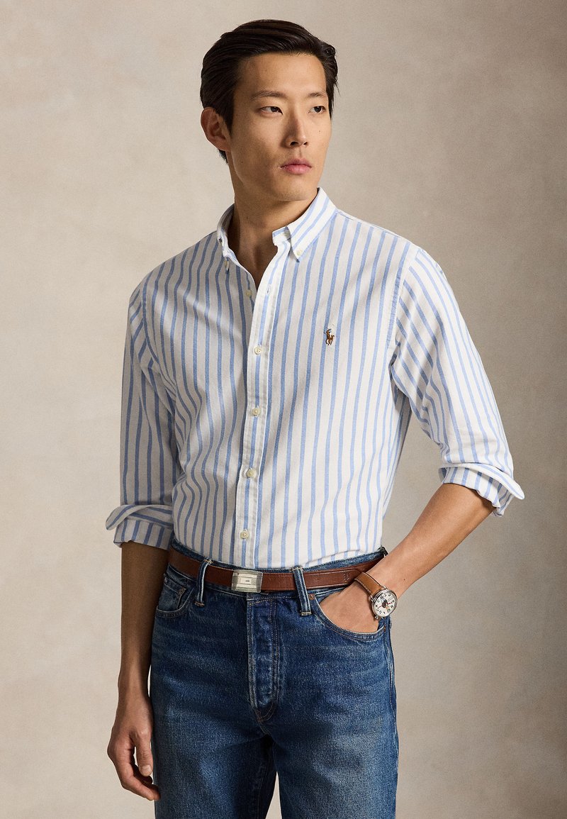 Mann trägt ein weißes und blau gestreiftes Hemd mit Knöpfen und aufgekrempelten Ärmeln, blaue Jeans, einen braunen Gürtel und eine Armbanduhr, steht mit einer Hand in der Tasche.