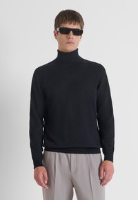 Maglione a collo alto nero in materiale morbido, design aderente con maniche lunghe. Modello abbinato a occhiali da sole neri e pantaloni grigi chiari.
