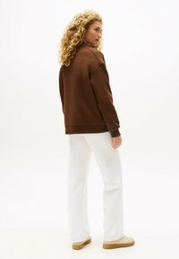 Bruine oversized sweatshirt met geribbelde manchetten, gecombineerd met witte broek met wijde pijpen en lichtgekleurde sneakers. Kruidachtige haren zijn zichtbaar van achteren.