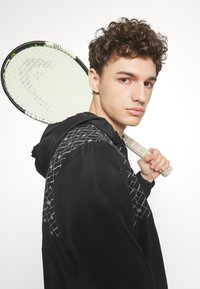 Svart huva jacka med geometriskt vitt mönster, bärs av en person som håller en tennisracket med ett gult snömmönster.