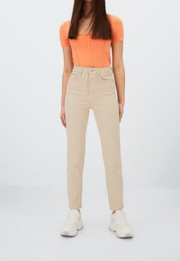Pantalones beige de talle alto con un corte ajustado, fabricados en algodón, que cuentan con bolsillos frontales y cierre de botón, combinados con una blusa naranja ajustada y zapatillas blancas.