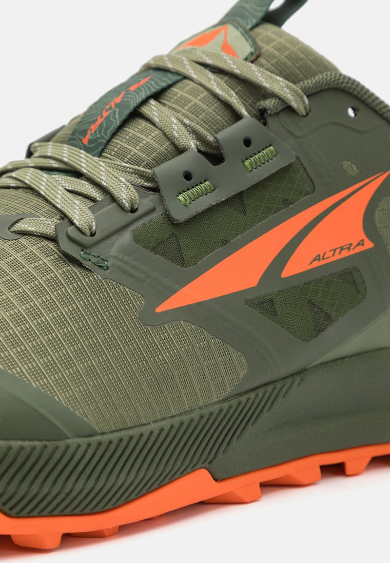 Zapatilla deportiva verde con parte superior texturizada, detalles en naranja y una suela robusta con tacos prominentes para un buen agarre.