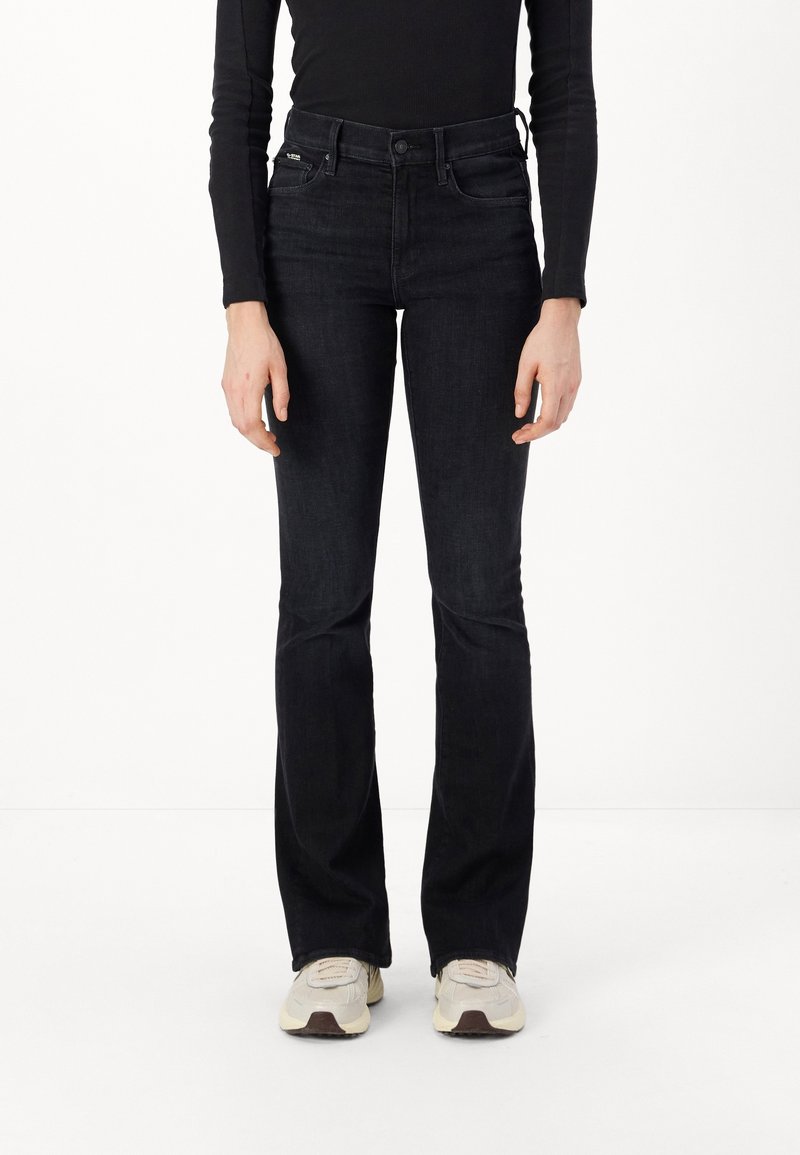 Jeans de mezclilla oscura de corte acampanado con una cintura alta, costuras visibles, dos bolsillos delanteros y una textura suave, combinados con unas zapatillas de color claro.