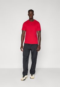 Rød T-skjorte med korte ermer og svart logo, kombinert med svarte, løstsittende bukser og beige sneakers, stående mot en ensfarget grå bakgrunn.