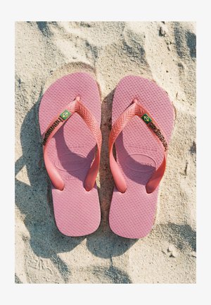 Paire de tongs Havaianas roses avec des icônes du drapeau brésilien reposant sur une surface de plage sableuse claire.