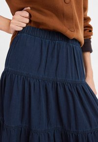Gonna a strati blu navy realizzata in tessuto leggero, con una vita elastica e un orlo arricciato, abbinata a un maglione marrone.