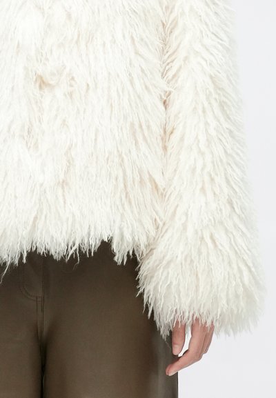 Plyšová biela skrátená bunda s dlhými, nadýchanými rukávmi. Vyrobená z mäkkého materiálu, má textúrovaný, huňatý vzhľad. Kombinovaná s hnedými nohavicami.