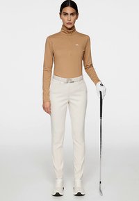 Beżowy golf z długim rękawem z suwakiem przy kołnierzu, o fakturowanym wzorze. Kremowe spodnie z wąską nogawką oraz kij golfowy, noszone z białą rękawiczką.