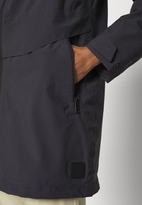 Musta vedenpitävä takki, jossa on vetoketjullinen tasku, nepparikiinnikkeet hihansuissa ja logopahvi lähellä helmaa. Suora pinta ja moderni leikkaus.