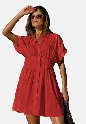 Žena v červených šatech s krátkými rukávy s výšivkou, držící brýle na slunce v jedné ruce a v druhé tmavý tkaný sáček.