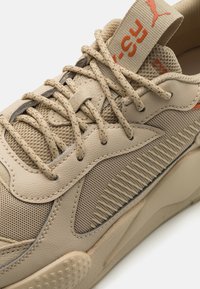 Baskets de sport beiges avec un mesh texturé et des panneaux synthétiques lisses, présentant un design à motifs et des accents orange contrastants sur la languette.