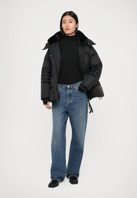 Jachetă puffer neagră cu guler din blană artificială, asortată cu blugi largi de culoare albastru deschis. Materialul este neted, cu un finisaj mat.
