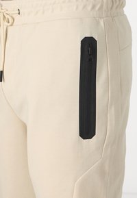 Beige Jogginghose mit strukturiertem Finish, ausgestattet mit einer schwarzen Reißverschlusstasche an der Seite und einem Kordelzugbund für eine verstellbare Passform.