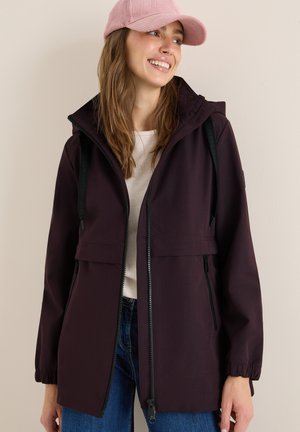 Veste imperméable violette avec capuche, poignets élastiques et fermeture éclair à l'avant. Portée par-dessus une légère chemise, associée à un jean bleu.