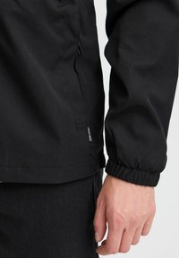 Jachetă neagră dintr-un material neted și ușor, cu un buzunar cu fermoar și manșete elastice. O etichetă cu logo este vizibilă pe tivul de jos.