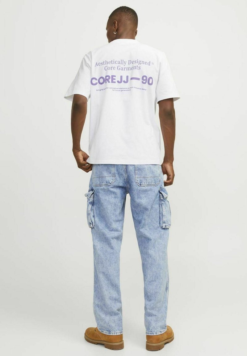 Hvid bomulds-T-shirt med lilla tekst på ryggen, der står "Æstetisk designede kerne beklædning COREJJ-90," parret med lys blå cargo bukser.