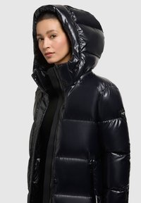 Padded černá bunda s lesklým povrchem, oversize kapucí, zipovým zapínáním a elastickými manžetami. Obsahuje logo na levém rukávu a zipové kapsy.