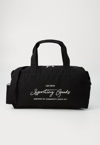 Bolsa de lona negra tipo duffel con asas dobles, correa de hombro desmontable y texto impreso en blanco que presenta el nombre y lema de la marca.