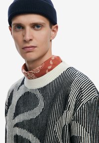 Stickad tröja med svartvita ränder och grafisk design, hög rundad krage och en kontrasterande bandana i rostfärg.