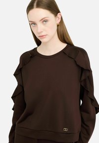 Brązowy sweatshirt z warstwowym efektem falbanek na rękawach, strukturalnym ribem oraz małym złotym logo blisko dołu.