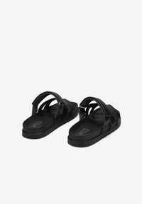 Black sandals with a flat sole, featuring dual straps. The material is smooth with a slight sheen. Includes a logo detail on the footbed.