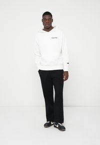 Junger Mann steht, trägt einen weißen Kapuzenpullover mit Logo, schwarze Hose und schwarz-weiße Turnschuhe vor einem schlichten weißen Hintergrund.