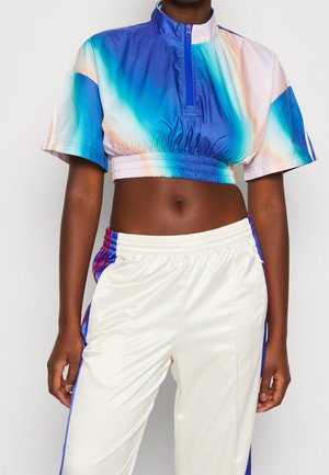 Personne portant un haut court à manches courtes avec un dégradé bleu et blanc, une fermeture éclair, et un pantalon de sport blanc avec des bandes bleues et rouges.