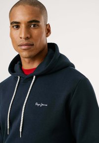 Sudadera azul marino de algodón, con un bolsillo canguro en la parte delantera, cordones crema y un logo bordado en blanco en el pecho.