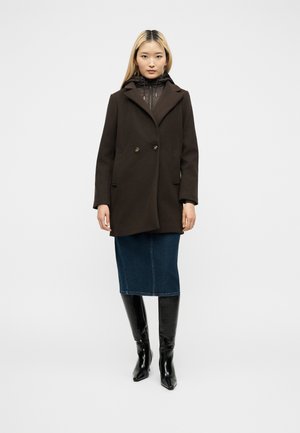 Femme asiatique debout portant un manteau marron foncé par-dessus un sweat à capuche noir, une jupe en jean bleue et des bottes noires montant jusqu'aux genoux sur fond blanc.