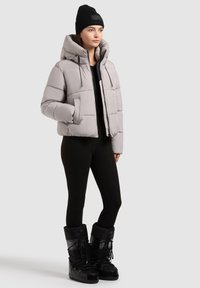 Giacca puffer grigia con cappuccio e cerniera frontale, dotata di lacci elastici e tasche laterali. Indossata sopra un top nero e leggings, abbinata a stivali neri.