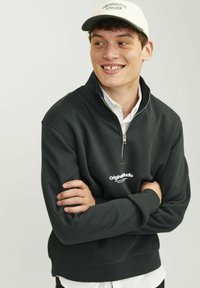 Sudadera de cremallera en gris oscuro con un cuello alto, que presenta un logo blanco. Llevada sobre una camisa blanca, con las manos cruzadas al frente.