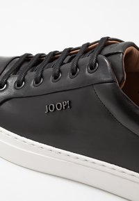 Baskets en cuir noir avec lacets noirs, œillets argentés et semelle blanche. Présentent un logo "JOOP!" en relief sur le côté. Texture lisse.