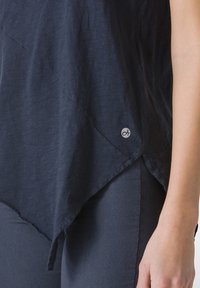 Top asimetric de culoare albastru marin, realizat dintr-un material ușor, cu o margine brută și un mic logo pe lateral. Asortat cu pantaloni închiși la culoare.
