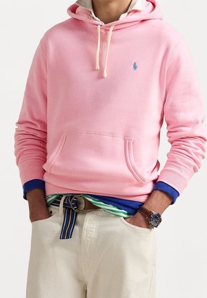 Persona que lleva una sudadera con capucha rosa claro con logo azul, sobre camisas de rayas azules y verdes, pantalones beige con un cinturón de tela a rayas y un reloj de pulsera.