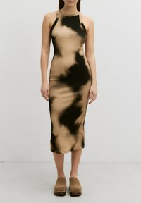 Figurbetontes, ärmelloses Kleid in einer Mischung aus beigen und schwarzen Batikmustern. Aus einem glatten Stoff gefertigt, knielang, kombiniert mit braunen Holzschuhen.