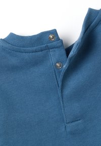 Dettaglio della camicia di cotone blu con colletto a coste, chiusura con bottoni a pressione e texture liscia. Presenta una fessura laterale e orlo cucito.