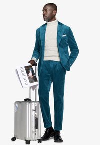 Teal velour-puku, jossa on teksturoitu valkoinen poolopaita. Pitelee aikakauslehteä ja seisoo hopeisen vetolaukun vieressä. Mustat kengät.