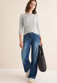 Světlé šedé žebrované tričko s dlouhým rukávem je kombinováno s modrými kalhotami s širokými nohavicemi. Modelka drží černou péřovou kabelku a má na sobě bílé tenisky.