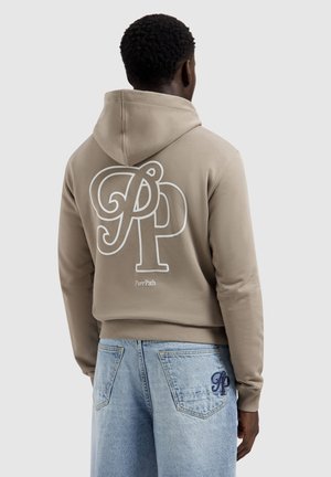Man wearing beige hoodie with large "PP" logo and "Pure Path" text on back, paired with light blue jeans embroidered with "PP" on pocket.