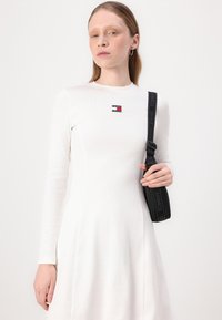 White long-sleeve dress with a fitted bodice and flared skirt, featuring a small red, white, and blue logo patch. Black strap bag.